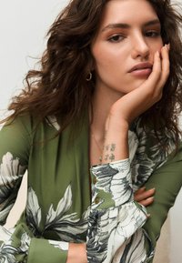 Žalia gėlių rašto palaidinė ilgomis rankovėmis, su raštuotais rankogaliais ir sagutėmis. Modelis turi natūralų makiažą ir remiasi smakru ant rankos.