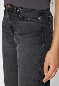 Juodi džinsai slim fit stiliaus, su variniais mygtukais ir vinimis, priekinėmis kišenėmis ir subtiliu plaukimo tekstūros akcentu audinyje.