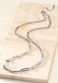 Silver chain necklace featuring elongated links interspersed with round spacers, displayed on a light stone surface. Long and flexible design.