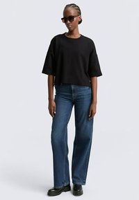 Schwarzes, kurz geschnittenes T-Shirt mit weiten Ärmeln, kombiniert mit hoch taillierten blauen Jeans. Die Person trägt eine schwarze Sonnenbrille und klobige schwarze Stiefel.