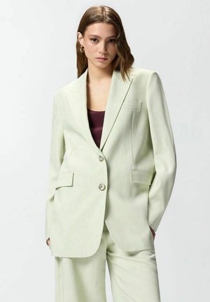 Jonge vrouw die een lichtgroed, op maat gemaakt pak draagt met brede revers en twee knopen, gecombineerd met een donkerpaarse top en gouden oorringen.