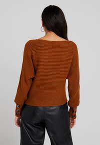 Brauner gerippter Strickpullover mit weitem Ausschnitt und langen Ärmeln, mit Knopfdetails an den Bündchen, kombiniert mit schwarzen Lederhosen.