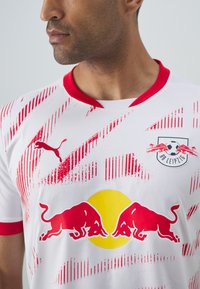 Balta futbolo marškinėliai su raudonais akcentais ir įstrižais raštais, su raudonu bulių logotipu ir geltonu apskritimu, pagaminti iš lengvos medžiagos.