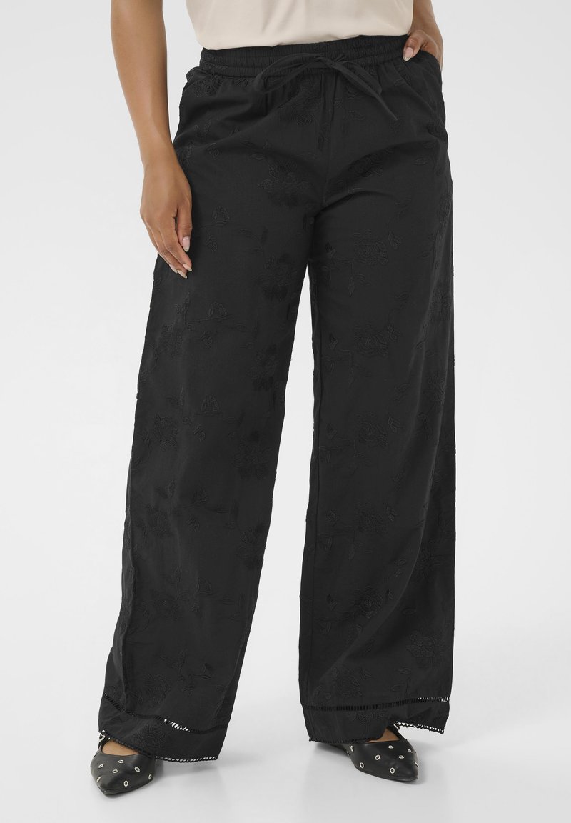 Laza fekete, virágmintás nadrág húzózsinóros derékrésszel, fekete hegyes orrú lapos cipőkkel, ezüstszínű fémfülekkel viselve.