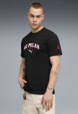 Jonge man met kort haar die een zwart AC Milan Puma T-shirt draagt, beige broek en tatoeages op zijn onderarmen, staand voor een grijze achtergrond.