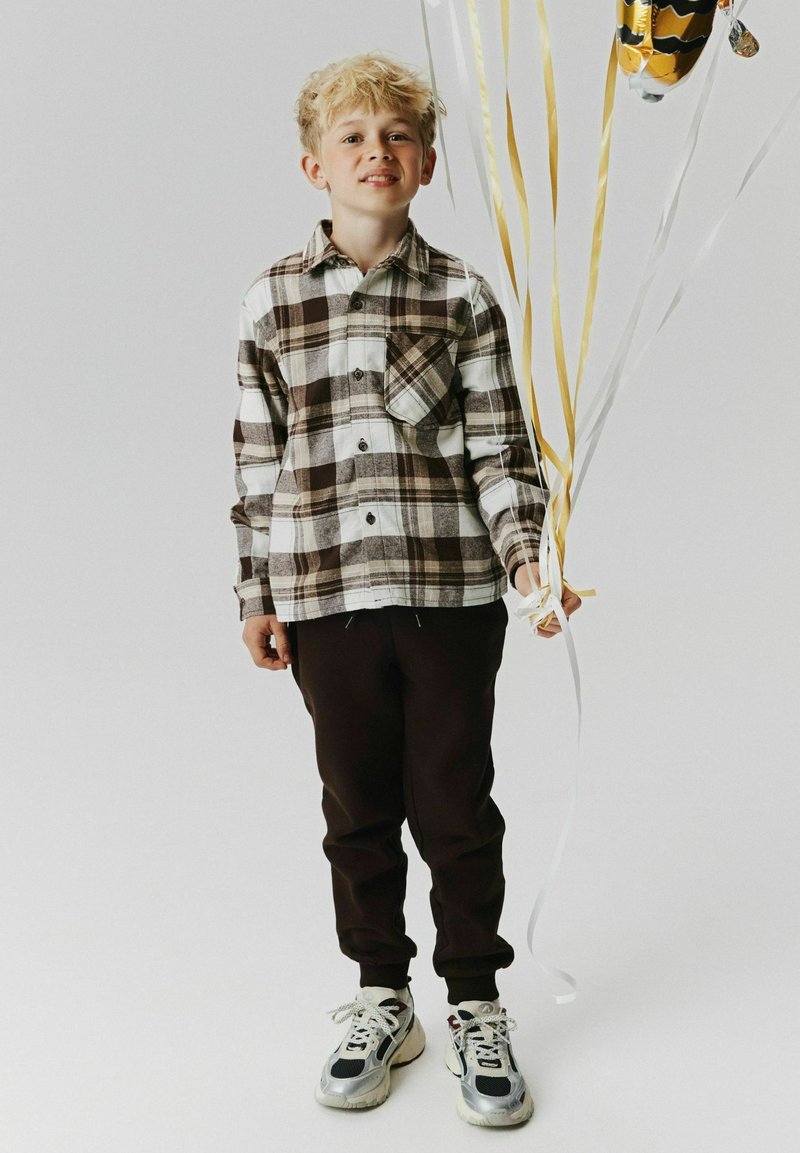 Garçon aux cheveux blonds portant une chemise à carreaux et un pantalon foncé, tenant des ballons dorés et blancs sur un fond clair uni.