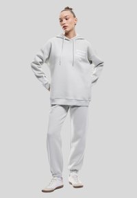 Conjunto de sudadera y pantalones de chándal en gris claro, hecho de tejido suave. La sudadera cuenta con un bolsillo frontal y un detalle de texto en el pecho. Zapatillas blancas completan el look.