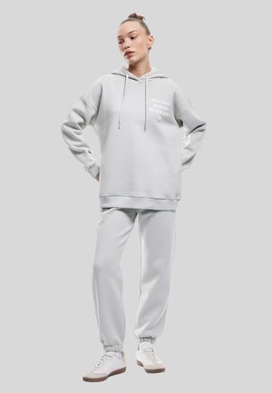 Conjunto de sudadera y pantalones de chándal en gris claro, hecho de tejido suave. La sudadera cuenta con un bolsillo frontal y un detalle de texto en el pecho. Zapatillas blancas completan el look.