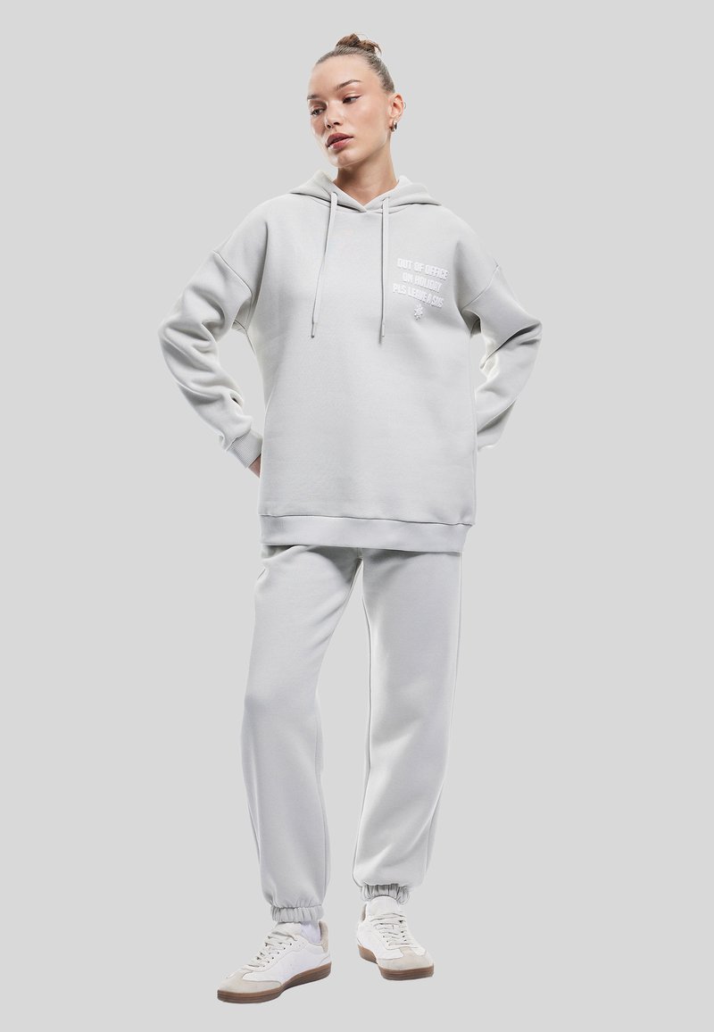 Conjunto de sudadera y pantalones de chándal en gris claro, hecho de tejido suave. La sudadera cuenta con un bolsillo frontal y un detalle de texto en el pecho. Zapatillas blancas completan el look.