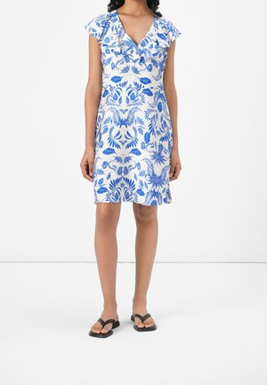 Kvinde iført hvid knælængde kjole med blåt blomsterprint og flæset V-hals, kombineret med sorte flade sandaler.