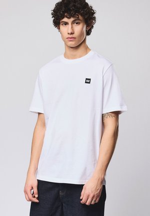 Giovane con capelli ricci che indossa una maglietta bianca semplice con un piccolo logo nero sul petto e pantaloni scuri su uno sfondo chiaro.