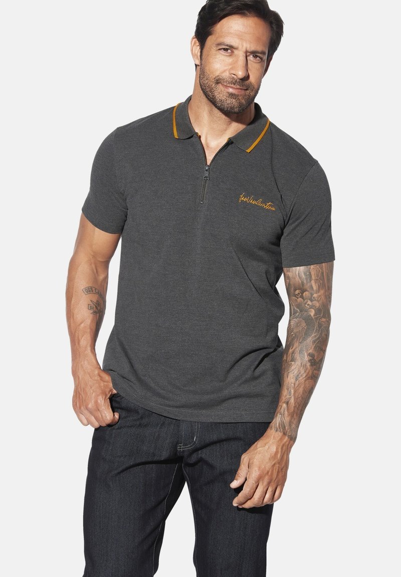 Dunkelgraues Polo-Shirt mit einem Reißverschlusskragen und kontrastierenden senfgelben Akzenten. Gesticktes Logo auf der Brust. Tragend mit dunklen Denim-Jeans.