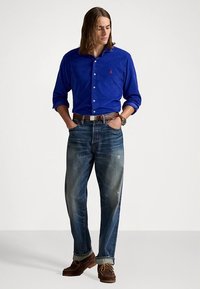 Kék gombos ing, világos farmernadrág kopott részletekkel, barna öv, barna fűzős cipő, hosszú haj. Egyszerű, hétköznapi öltözék.