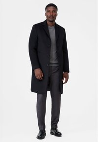 Svart ullrock med slagkragar, sex knappar och en smal grå tröja under. Kombinerad med grå skräddarsydda byxor och svarta läderstövlar.