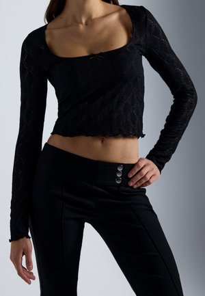 Femme portant un crop top noir à manches longues avec motif losange texturé et un pantalon noir taille haute avec trois boutons.