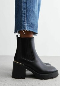 Stivali neri in pelle alla caviglia con pannelli elastici rigati, tacco spesso nero e accentatura dorata alla base. Indossati con jeans blu strappati.
