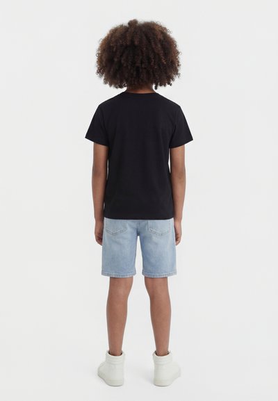 Copil cu păr creț, purtând un tricou negru, pantaloni scurți de denim bleu deschis și adidași albi, stând cu fața spre spate, pe un fundal simplu.