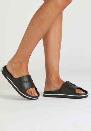 Ben iført sorte slid-sandaler med åbne tæer og hvide striber på sålerne, den ene fod løftet let fra jorden.