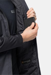 Sort, stribet jakke med en lynlås-lomme, der viser en hånd, der indsætter en smartphone. Funktioner inkluderer en glat tekstur og et logo-mærke for Microguard Ecosphere.