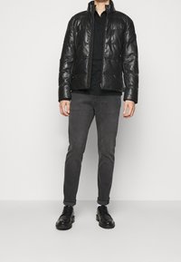 Veste matelassée noire avec un col montant, une fermeture éclair à l'avant et une texture brillante, associée à un pantalon chino gris foncé et des chaussures noires.