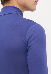 Kratak plan osobe koja nosi usku plavu majicu dugih rukava, prikazujući rame, vrat i djelomični bočni profil nasuprot jednobojnoj pozadini.