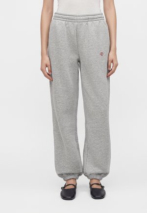 Persona che indossa pantaloni della tuta grigio chiaro con polsini elastici, piccolo logo rosso e ballerine nere, in piedi davanti a uno sfondo bianco semplice.
