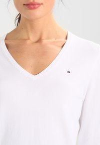 Nainen valkoisessa V-kaula-aukkoinen villapaidassa, jossa on pieni punaisen, valkoisen ja sinisen logo rinnassa, kuvattuna olkapäistä leukaan.