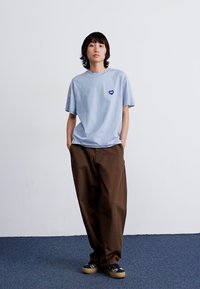 Svijetloplavi majica kratkih rukava s malim ljubičastim srcem na prsima, u kombinaciji s smešnim širokim hlačama i šarenim obućom.