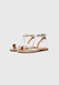 Metallische zilveren platte sandalen met een minimalistisch ontwerp, voorzien van twee brede banden, een enkelband met kettingdetail en een tan kleurige zool.