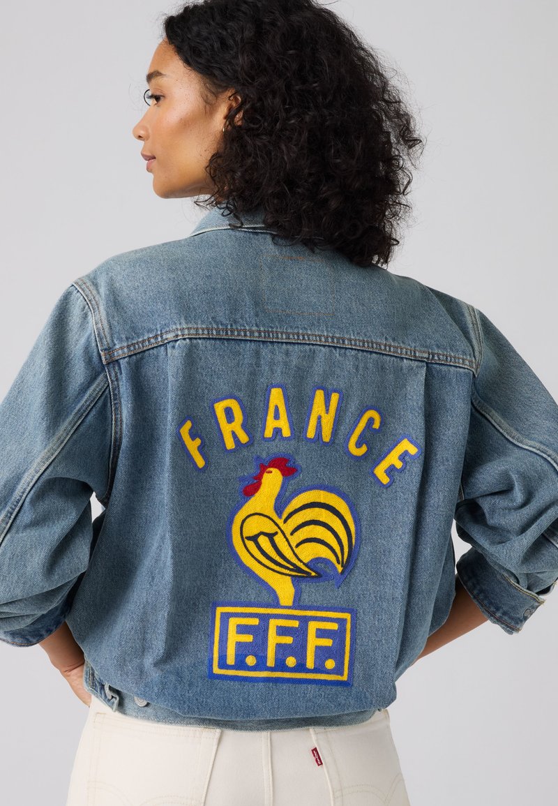 Donna con capelli ricci che indossa una giacca di jeans con un emblema giallo di gallo e la scritta "FRANCE F.F.F." sulla schiena.