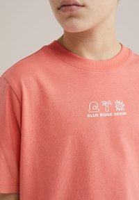 Coralowa bawełniana koszulka z okrągłym dekoltem. Z przodu znajduje się biały napis i ikony z napisem "BLUE RIDGE DENIM". Miękka w dotyku.