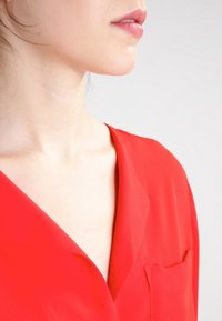 Blusa rossa con scollo a V, tessuto morbido e una tasca sul petto. Texture liscia e vestibilità fluida, che mette in evidenza il colletto e l'area delle spalle.