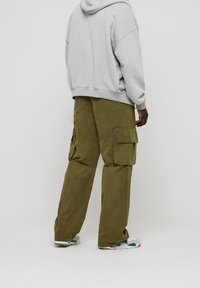 Pantalon cargo olive avec plusieurs poches latérales, en coton et à coupe ample. Associé à un sweat à capuche gris et des baskets blanches avec des accents verts.