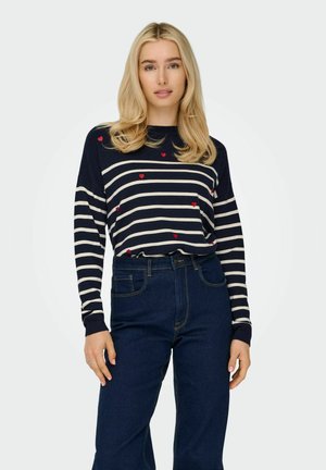 Marineblauw en witte gestreepte langemouwen top met rode hartaccenten; gecombineerd met high-waisted donkerblauwe jeans; ribgebreide boorden tonen textuur.