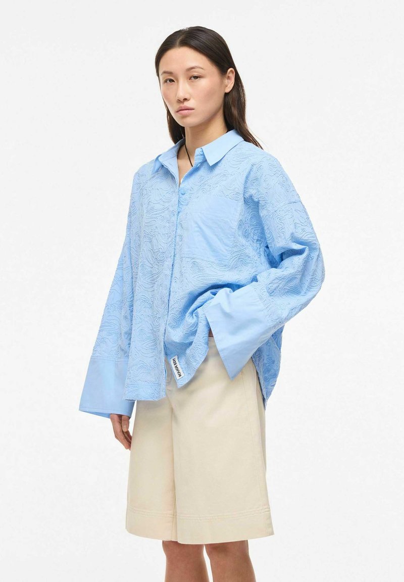 Mujer con camisa azul clara de gran tamaño con estampado y puños anchos y pantalones cortos beige hasta la rodilla, posando frente a un fondo blanco.