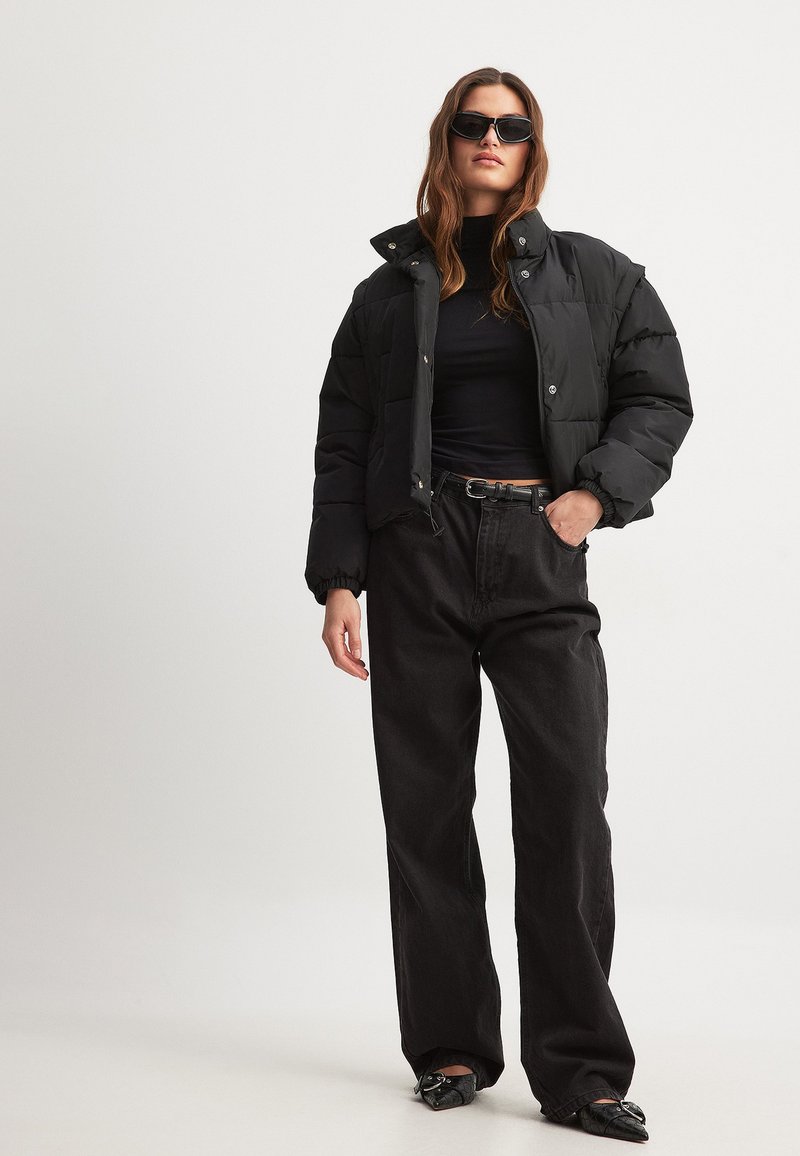 Must musternööriga must puffjakker, millel on lühendatud disain, kõrge krae ja nööbid, koos kõrge vöökohaga mustade teksade ja teravakas eluga kingadega.