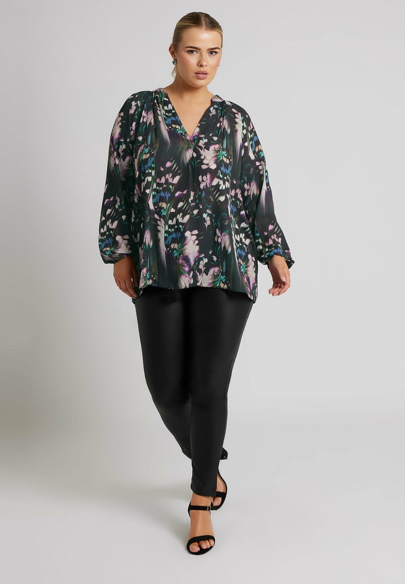 Blusa floreale nei toni del nero, verde e viola, con maniche lunghe e scollatura a V, abbinata a leggings neri aderenti e sandali neri con cinturini.