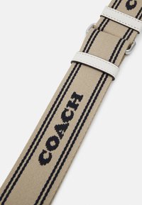 Bracelet de montre en tissu beige avec texte "COACH" noir et deux bandes parallèles noires, doté de boucles en cuir blanc et d'une boucle en métal argenté.