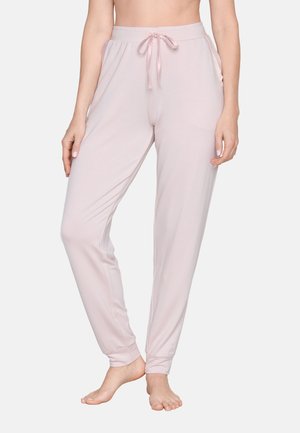 Donna che indossa pantaloni da lounge rosa chiaro con coulisse, in piedi a piedi nudi su uno sfondo semplice.