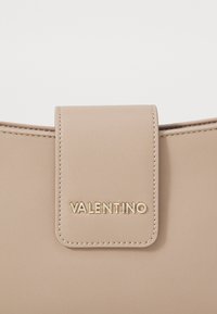 Μπεζ δερμάτινη τσάντα χειρός με μαγνητική κλείσιμο καπάκι, διακοσμημένη με λογότυπο "Valentino" σε χρυσό χρώμα και προσεγμένη ραφή γύρω από τις άκρες.