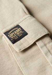 Beige stof met een zwart en geel Superdry Work-Wear label gestikt op een opgevouwen zakflap.