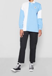 Ljusblå och vit långärmad pikétröja med krage, med en rund logotyp. Kombinerad med svarta cargo-byxor och svarta högstövlar.