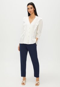 Blusa bianca con pieghe verticali e dettagli di rouches lungo il collo, con bottoni dorati. Abbinata a pantaloni blu navy a sigaretta.