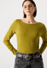 Langermet, olivengrønn topp med bred U-hals, kombinert med høytlivede svarte jeans og et naglet svart belte. Tilbehør inkluderer sølvøreringer og store solbriller.