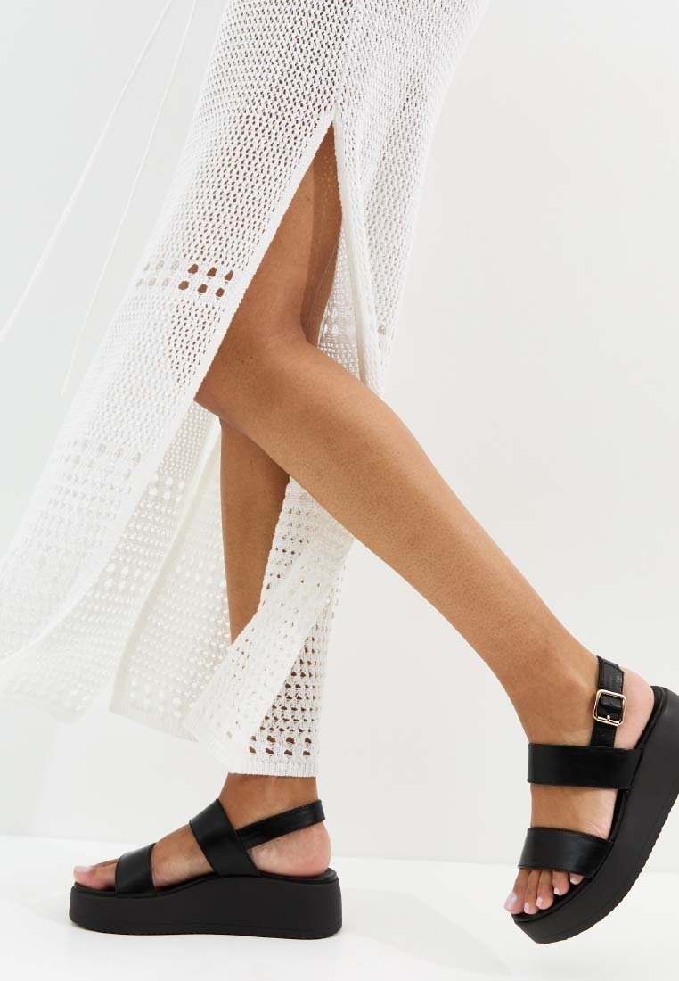 Piernas con sandalias negras de plataforma con tiras en el tobillo, combinadas con un vestido de punto blanco con aberturas laterales.