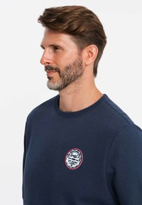 Sudadera azul marino hecha de un tejido suave, con cuello redondo y un logotipo bordado circular en el pecho con acentos en rojo y blanco.