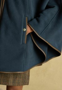 Ruka v vrecku tmavomodrého fleecového plášťa s hnedým lemovaním, pod ktorým je vidieť károvanú sukňu na neutrálnom pozadí.