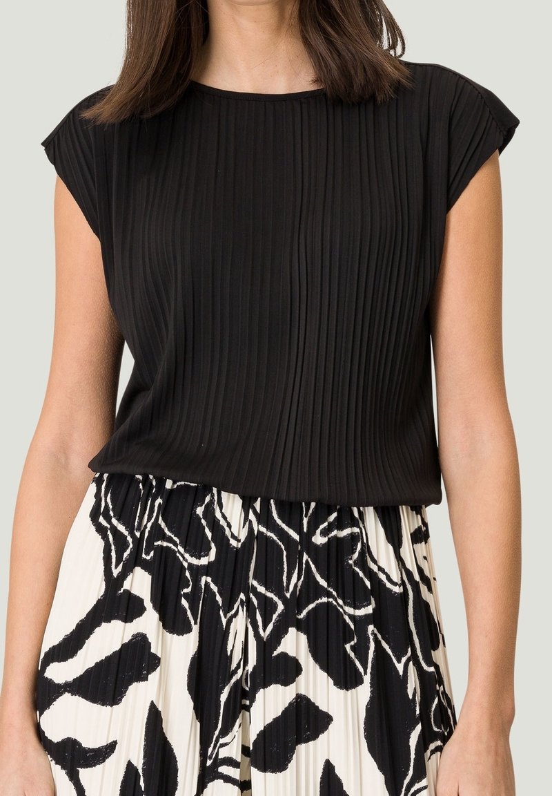 Femme portant un haut noir plissé à manches courtes et une jupe plissée taille haute avec un motif abstrait noir et blanc.