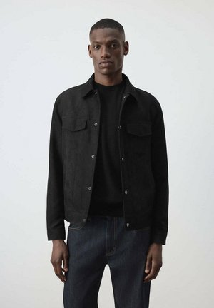 Hombre con el pelo corto vestido con una chaqueta negra abotonada sobre una camiseta negra y vaqueros azul oscuro, de pie contra un fondo claro y liso.