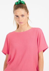Blusa rosa a maniche corte con un tessuto testurizzato e leggermente sgualcito. Girocollo con vestibilità rilassata. Fascetta per capelli verde visibile nei capelli.